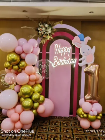 I created this adorable setup for a little one's first birthday. It features a pink and gold balloon arrangement, a custom 'Happy Birthday' neon sign on a pink arched backdrop, a cute rabbit character, and a large rose gold number '1' foil balloon.