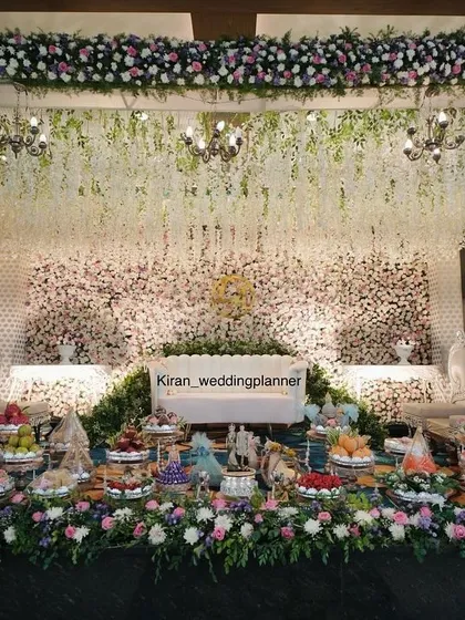 An elegant engagement stage with a backdrop of white flowers and hanging chandeliers. The foreground is beautifully arranged with traditional sweets and gifts, all framed by lush floral borders.