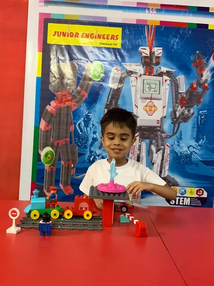 A happy student shows off his completed LEGO Duplo train set project in front of a robotics-themed poster.