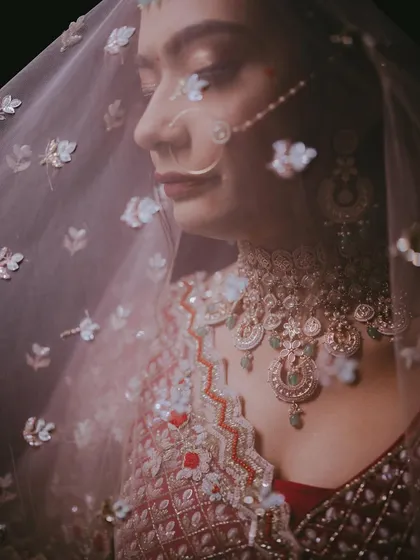 A close-up shot through the veil, focusing on the bride's profile, her elegant nose ring, and the intricate details of her necklace.