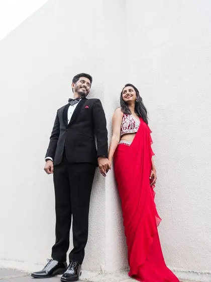 A modern and stylish couple pose against a minimalist white wall. The bold red saree and classic tuxedo create a striking visual contrast, perfect for a contemporary pre-wedding shoot.