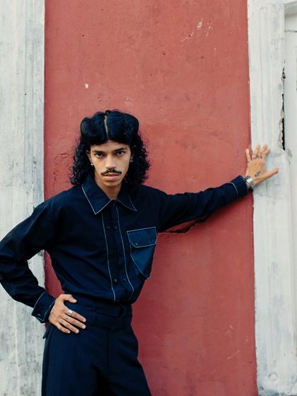 A confident pose against a red and white wall. The composition uses strong vertical lines to complement the model's stance, creating a powerful and graphic fashion photograph.