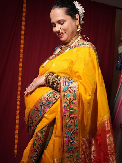 This image shows a close-up of the intricate border of a Paithani saree, a popular choice for traditional maternity shoots. The focus is on the rich textile and the mother's hands on her belly.