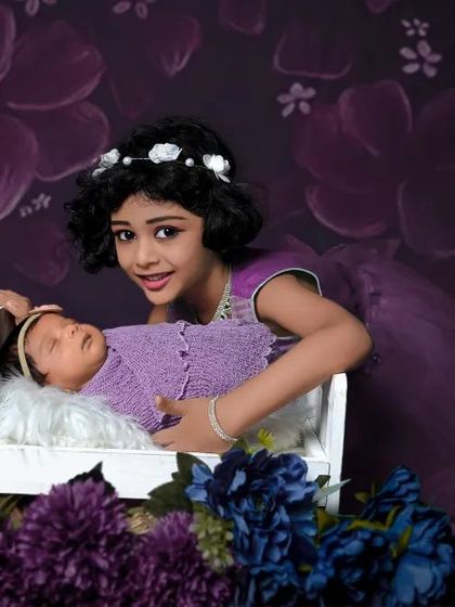An older sister proudly stands by her newborn sibling's crib. The coordinated purple outfits and floral background make this a beautiful sibling portrait.