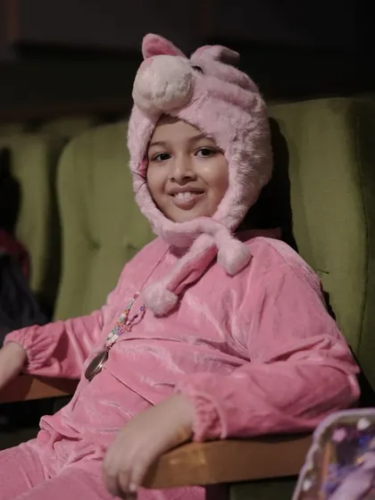 A young actor in a pig costume, patiently waiting in the audience seats before his scene. This shows the discipline and patience they learn.