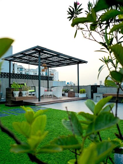 A view through the greenery of a simple yet elegant black metal pergola. It creates a defined, raised seating deck on the terrace, separating the lounge from the lawn area.