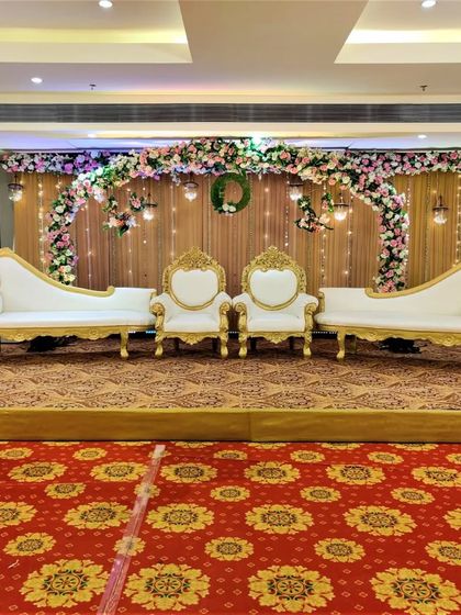 A rustic-themed stage decoration with a wooden texture backdrop and a floral arch. The traditional gold and white seating provides a beautiful contrast, perfect for an engagement ceremony.