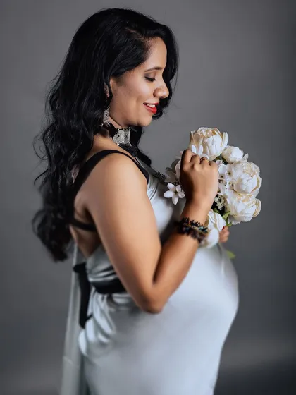 A close-up profile shot of a smiling mother-to-be in a silver saree, holding a bouquet of white flowers. The focus is on her happy expression and the gentle way she holds the bouquet.
