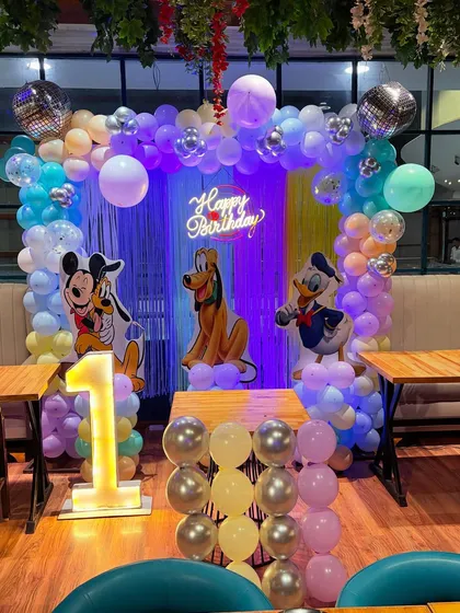 A classic Mickey Mouse and friends theme for a first birthday. This setup includes character cutouts of Mickey, Pluto, and Donald Duck, a colorful balloon arch, and a light-up number '1'.