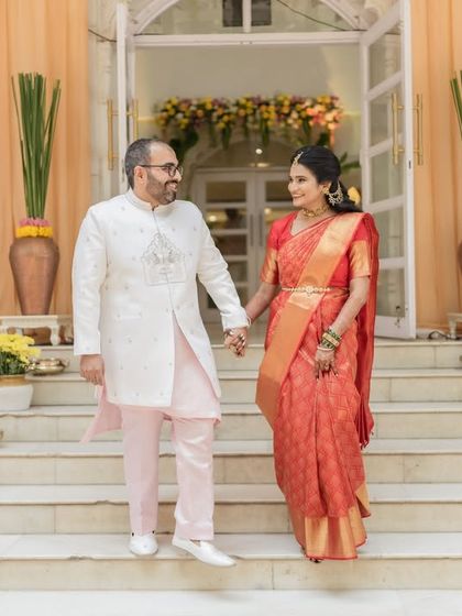 A full-length portrait of a couple holding hands on the steps of a beautifully decorated venue. A grand and elegant shot.