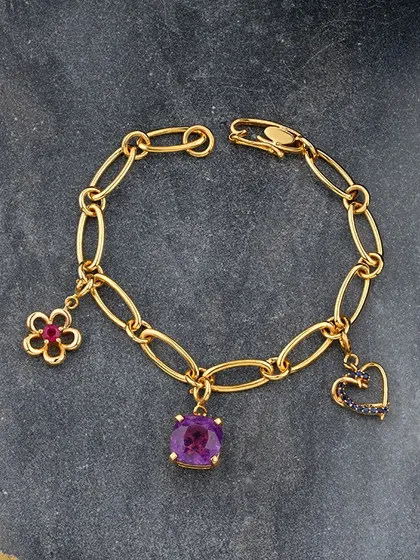 Charmed elegance. This stunning gold link bracelet is adorned with delicate charms: a ruby flower, a central amethyst, and a sapphire heart. It is perfect for adding a touch of personal luxury and colour to your everyday style.