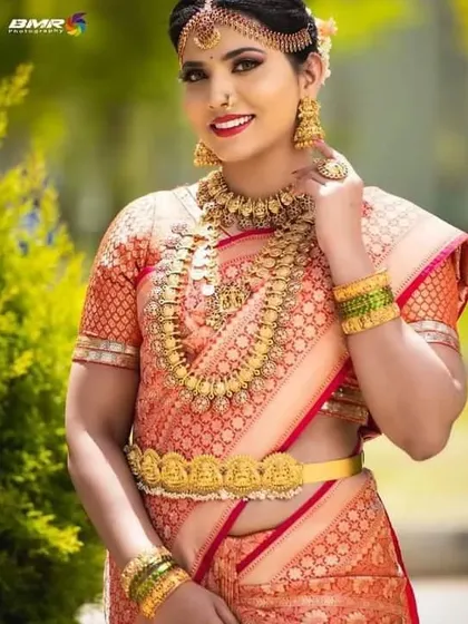 A radiant bride in an orange silk saree, adorned with our full coin necklace set and a simple gold-finish vaddanam.