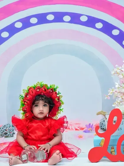 The baby girl sits amidst a colorful rainbow-themed set, complete with flowers and the word "Love". Her bright red dress stands out beautifully against the pastel colors.