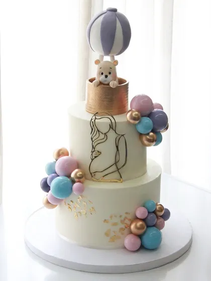 There's nothing sweeter than celebrating a new beginning. This two-tiered baby shower cake features a dreamy hot air balloon with a tiny teddy bear, all handcrafted from fondant, surrounded by a cascade of pastel chocolate spheres. It’s a gentle and elegant way to welcome a little one.