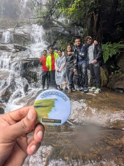 Our Netravathi trek badge held up against a backdrop of trekkers and a waterfall.