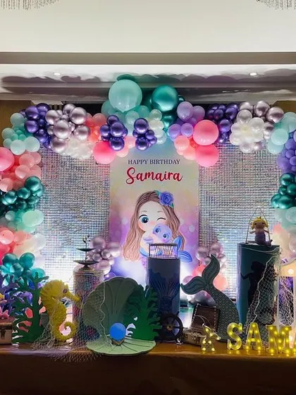 The main stage for the mermaid-themed party, featuring a vibrant balloon arch with chrome and pastel balloons. The setup includes a personalized backdrop, mermaid-themed props like seahorses and shells, and a light-up number '1'.
