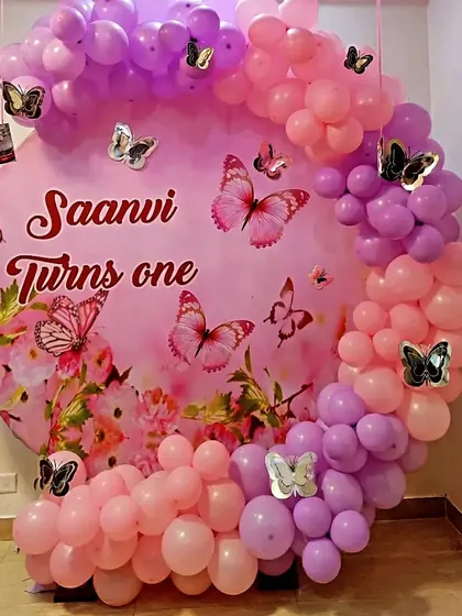 A lovely butterfly-themed first birthday decoration for "Saanvi". The design features a custom pink backdrop with the baby's name, surrounded by a beautiful arch of pink and purple balloons with decorative butterflies.