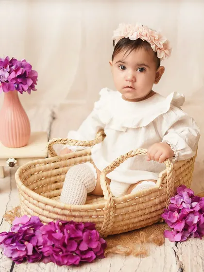 A baby girl in a simple white outfit, sitting in a wicker basket surrounded by purple flowers. The soft, rustic styling is perfect for a sitter session.