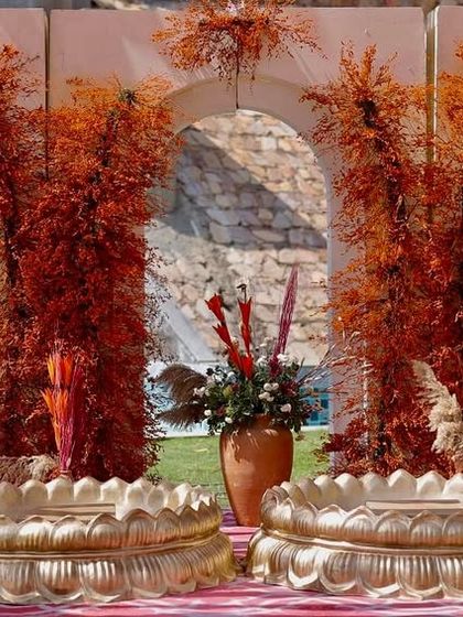 The main seating for the Haldi ceremony, featuring traditional brass urlis set against a backdrop of modern white arches adorned with fiery rust-orange foliage.