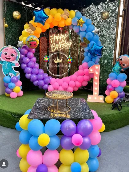 A vibrant Cocomelon first birthday party. This outdoor setup features a rainbow balloon ring, character cutouts, a "Happy Birthday" neon sign, and a light-up number '1'.
