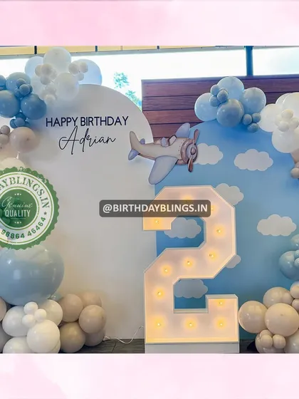 A dreamy airplane-themed second birthday party. The backdrop features a sky with fluffy clouds, and the balloons in shades of blue and white enhance the "up in the air" feeling.