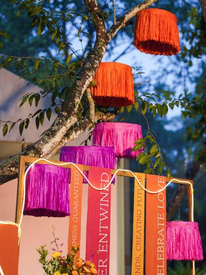 Colorful fringed lampshades hang from tree branches, adding a bohemian and festive touch to the outdoor Mehendi decor.