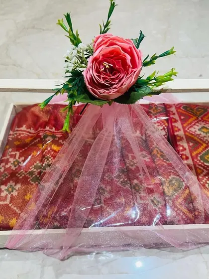 This is how I deliver a complete gift experience. A traditional Patola-style saree is carefully packed in a wooden tray with a clear acrylic top, wrapped in soft pink tulle, and finished with a floral arrangement, ready for any shagan ceremony.