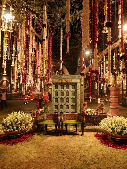 An enchanting outdoor setup under a tree adorned with hanging floral garlands. The antique-style door and traditional seating create a rustic yet magical atmosphere for an evening function.