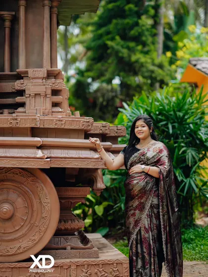 A solo portrait of an expectant mother in a shimmering saree, standing beside a stone chariot, creating a powerful and artistic image.