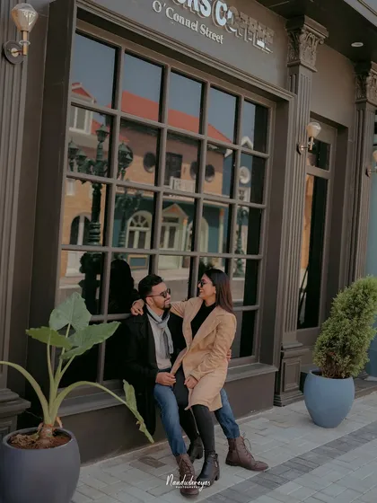 A couple sits together in front of a large window on a city street set, capturing a quiet, urban-cafe-style moment.