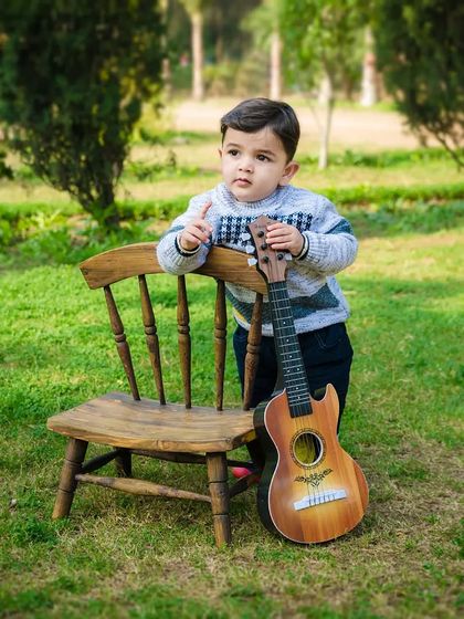 A toddler boy stands next to a small chair and a guitar, looking off into the distance. A cool and casual outdoor portrait.