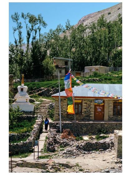A quiet corner of a Ladakhi village with a small stupa and prayer flags, a monk walking in the background.