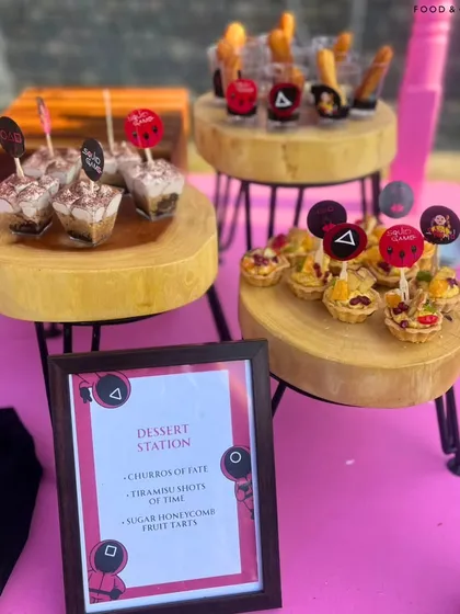 A close-up of the 'Squid Game' themed dessert station, featuring 'Churros of Fate' and 'Tiramisu Shots of Time'. The custom menu and themed toppers show how every detail is designed to fit your party's story.