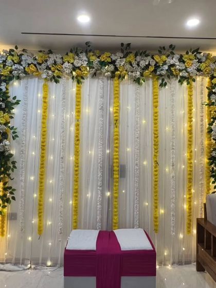 A simple and elegant backdrop for a home-based Haldi or pooja. The design uses white drapes with strings of fairy lights, framed by yellow and white flowers and marigold garlands.