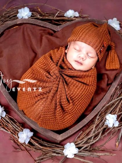 This rustic newborn portrait features a baby swaddled in a warm, russet-colored wrap, nestled inside a heart-shaped wooden bowl. The surrounding twig wreath adds a natural, earthy touch to the composition.