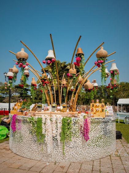 A stunning circular bar setup for an outdoor Mehendi. The mirrored base reflects the greenery, while the overhead structure with hanging pots and florals creates a beautiful focal point.