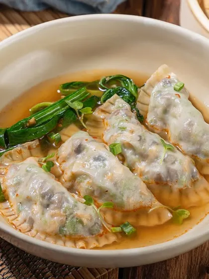 Gyoza-style dumplings in a clear broth. The natural styling with a bamboo mat and chopsticks gives an authentic feel.