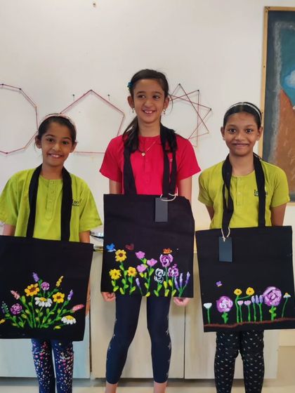 Three proud students from the school workshop displaying their unique floral designs painted on black tote bags. Each bag tells a different story through color and composition.