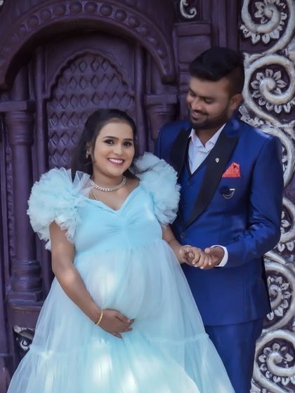 A stylish couple poses in front of an ornate door. The contrast between his sharp suit and her soft, flowing gown makes for a visually interesting and fashionable maternity photo.