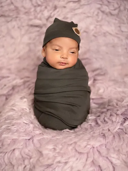 A newborn wrapped snugly in a dark grey swaddle against a soft lavender fur rug. The simple color palette creates a modern and serene portrait.