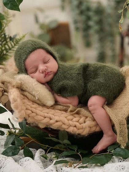 A newborn in a green outfit sleeps in a hanging basket adorned with vines, creating a beautiful and natural suspended portrait.