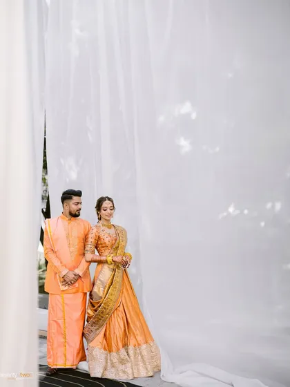 A full-length portrait of the couple in their coordinated orange outfits for the Haldi ceremony, standing against a simple white backdrop.