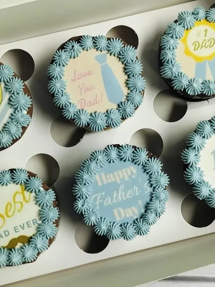 A full-box view of the pastel blue Father's Day cupcakes. The consistent light blue buttercream piping ties together the six unique designs, from a trophy to a simple "I Love Dad" message.
