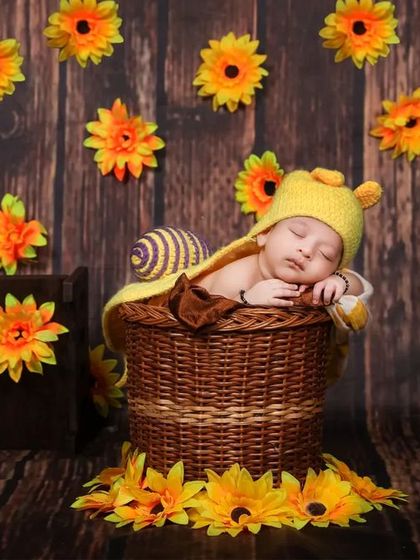 A whimsical setup featuring a baby dressed as a snail with a yellow knitted hat, nestled in a basket surrounded by sunflowers against a wooden backdrop.