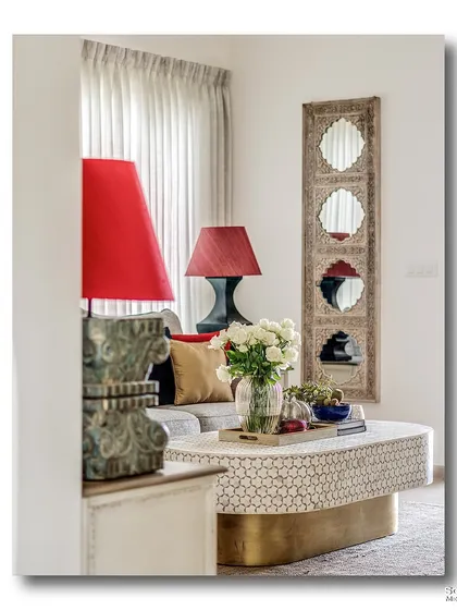 A peek into a living room, where two bold red lamps frame a comfortable sofa. The carved wooden mirror and patterned coffee table add layers of texture and traditional craftsmanship to the space.