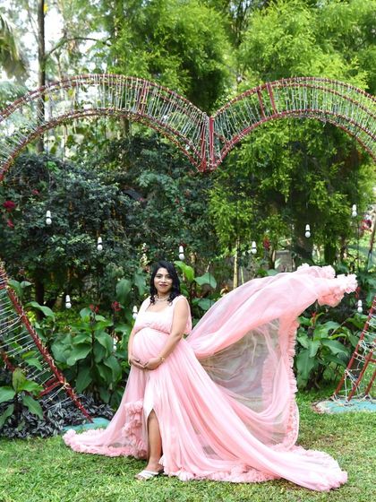 A dramatic outdoor shot featuring a mom-to-be in a flowing pink gown in front of a large heart-shaped arch. The fabric tossing in the wind creates a beautiful, dynamic effect.