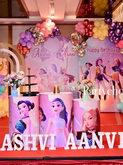 A vibrant K-Pop themed birthday party. The stage features a colorful backdrop with character art, themed pedestals, and a multi-colored balloon arrangement.