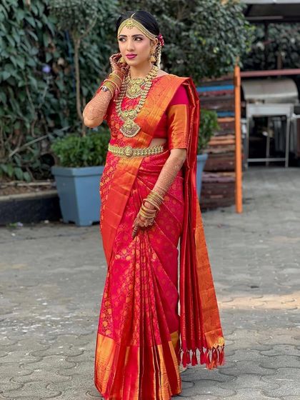 A full-length shot of a Tamil bride ready for her ceremony. The vibrant red and orange 9-yard saree is draped to perfection, showcasing the richness of the Kanjivaram silk.