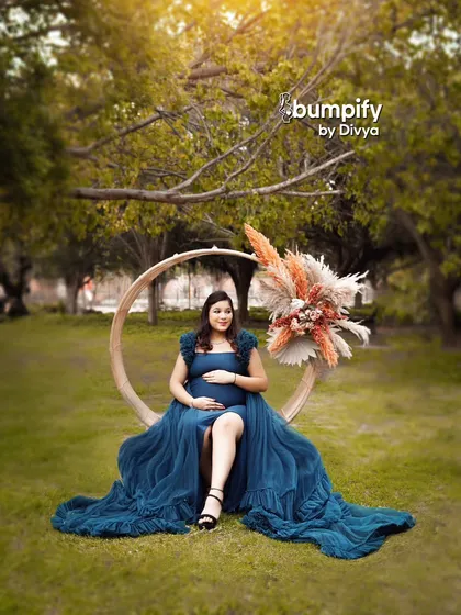 A striking outdoor portrait featuring a large wooden circle prop. The mom-to-be looks powerful and serene in a deep blue gown, seated within the circle.