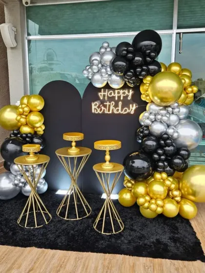 This outdoor terrace setup combines a black backdrop with a balloon arch in gold, silver, and black. The neon "Happy Birthday" sign and gold cake stands complete the look for a stylish evening celebration.